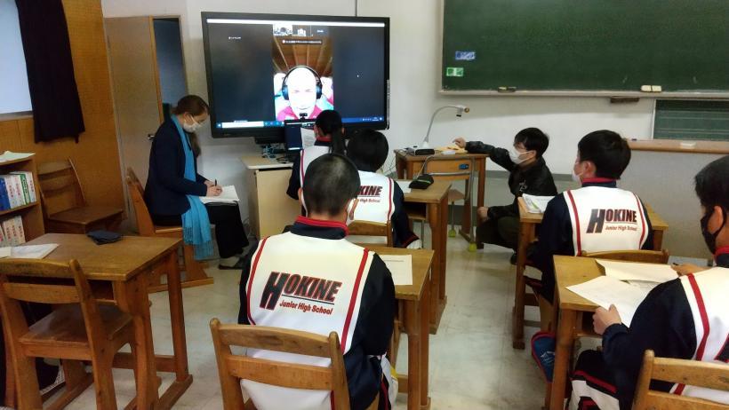 市立箒根中学校の教室でおこなわれたオンライン交流会にて、電子黒板の前にあるパソコンのカメラの高さに合わせて立膝でバッハマイヤー選手と会話する生徒を教室の後ろ側から撮影した写真