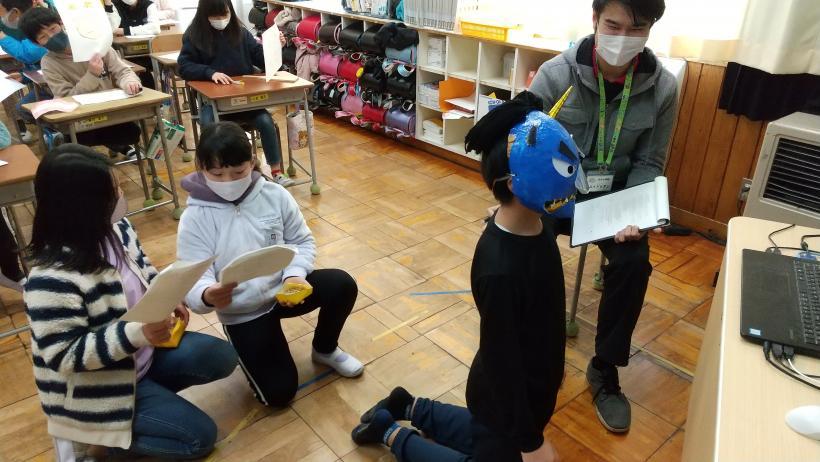 市立埼玉小学校の教室でおこなわれたオンライン交流会において、通信機材である電子黒板の前に、鬼の面をかぶって立膝になっている児童と、その後ろで小箱に入った豆を持ちながら屈んでいる児童2人の写真