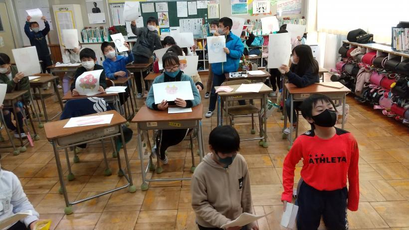 市立埼玉小学校の教室でおこなわれたオンライン交流会において、通信機材である電子黒板の前に座る児童2人と、その後ろの各自の席から鬼の顔を描いた絵をパソコンに見せる児童たちの写真