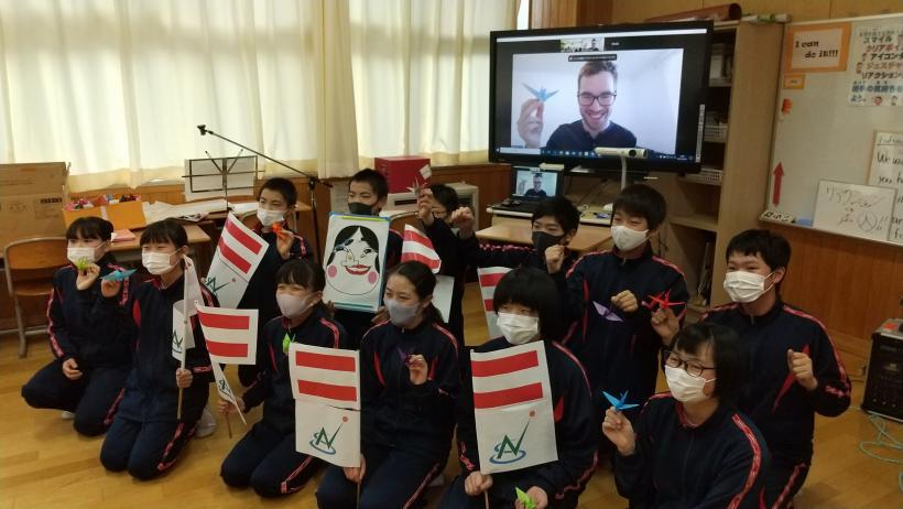 市立塩原小中学校の教室でおこなわれたオンライン交流会にて、電子黒板に映し出されたオネア選手とともに出来上がった福笑いやオーストリアの国旗を持って記念撮影をする7年生たちの写真