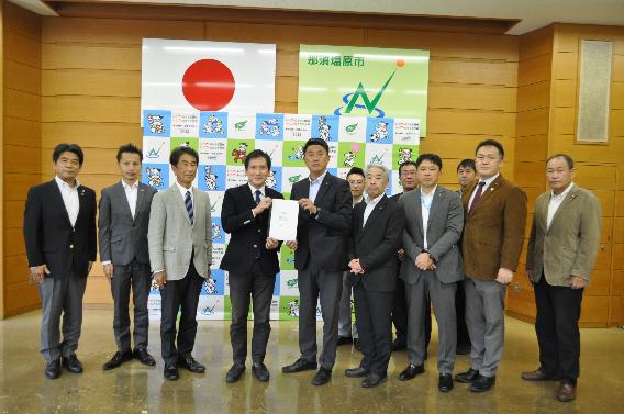 マスクをした大人が多数並んでいて中央の二人が提言書を手に持って掲げている写真