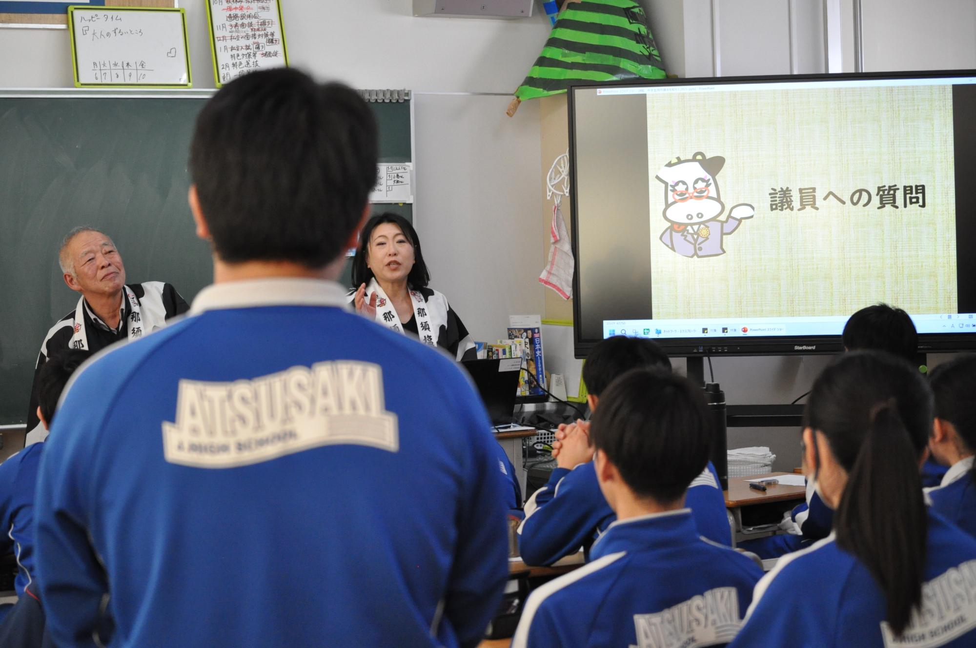 中学校の生徒が座っている前で、はっぴを着た男性と女性が話をしている写真